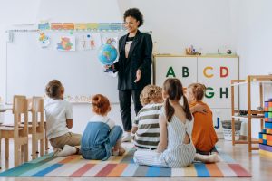 Teacher holding a globe talking to a circle of students in a decorated classroom. 