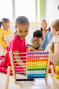 A young girl in a pink dress plays with a colorful abacus while other children and an adult are visible in the background, engaging in various activities.